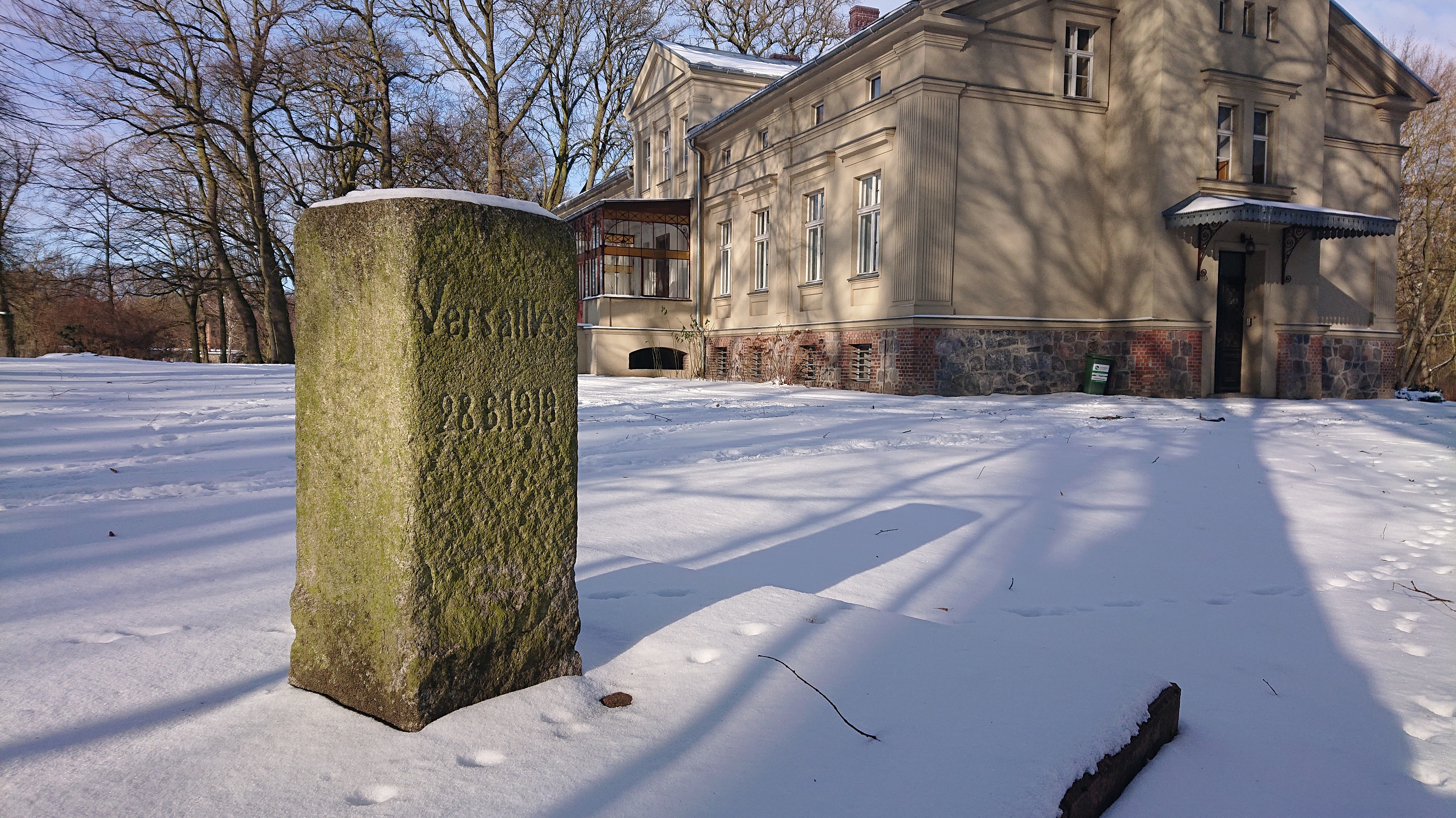 Kamienny słupek graniczny w śniegu na tle budynku dworu w Strychach.