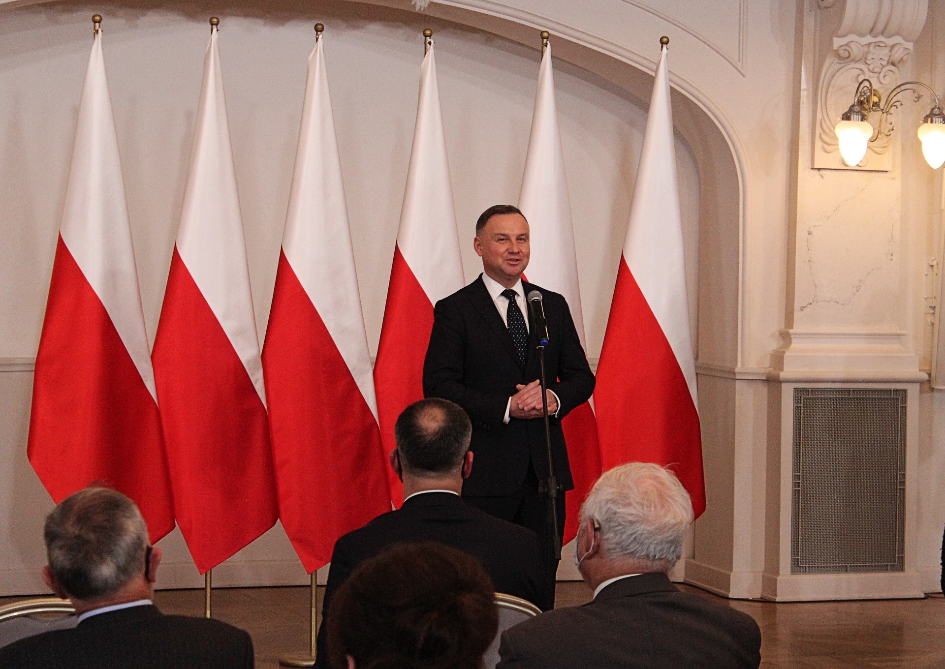 Prezydent RP Andrzej Duda przemawiający do zebranych słuchaczy na tle biało-czerwonych flag.