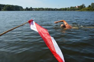 Zdjęcie przedstawia płynącą osobę w jeziorze, na pierwszy planie biało-czerwona flaga.