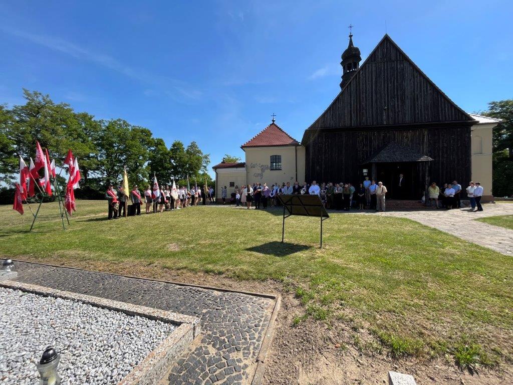 Od lewej flagi państwowe oraz powstańcze, dalej wszyscy uczestnicy uroczystości ustawieni na tle kościoła w Lgowie