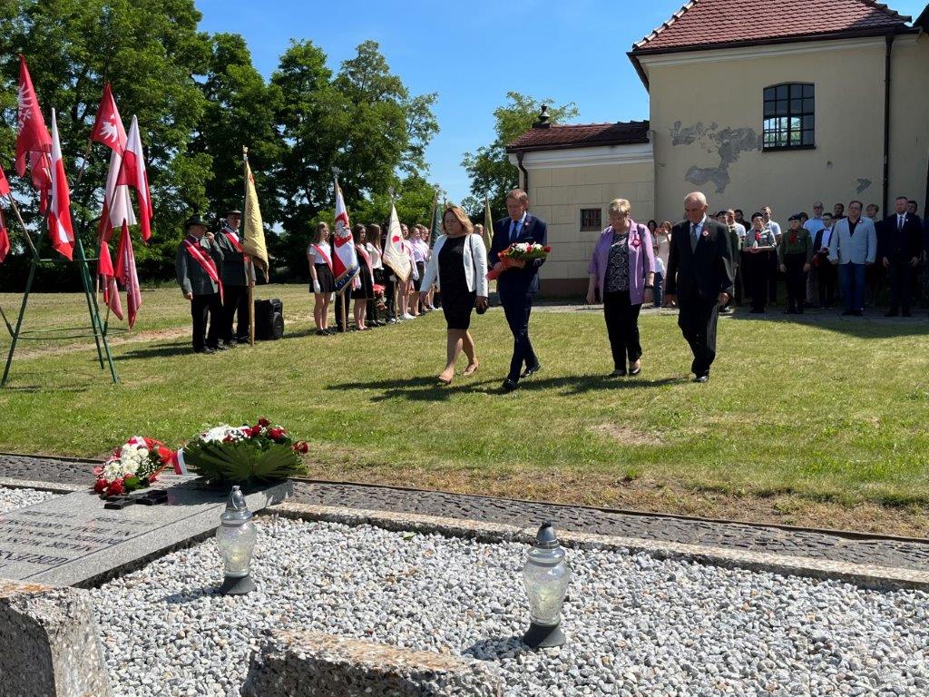 Delegacja władz lokalnych podchodzi do grobu majora z wiązanką kwiatów, w tle poczty sztandarowe oraz uczestnicy ceremonii.