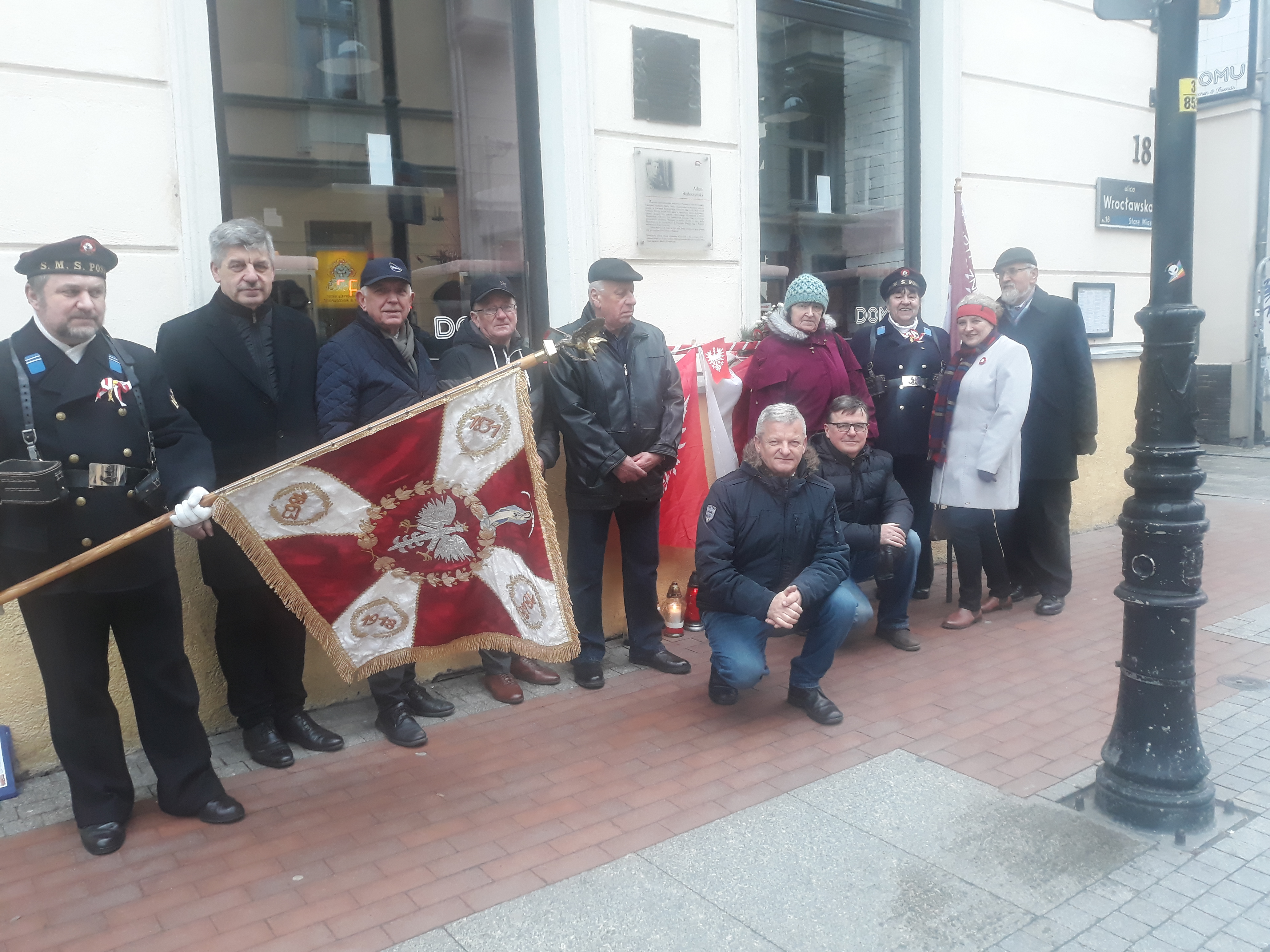 Na zdjęciu uczestnicy wydarzenia wraz ze sztandarem, ustawieni w rzędzie pod tablicą.