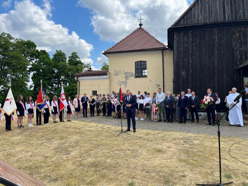 Mężczyzna przemawiający do mikrofonu, wokół stojące poczty sztandarowe. W tle widoczny fragment budynku kościelnego.