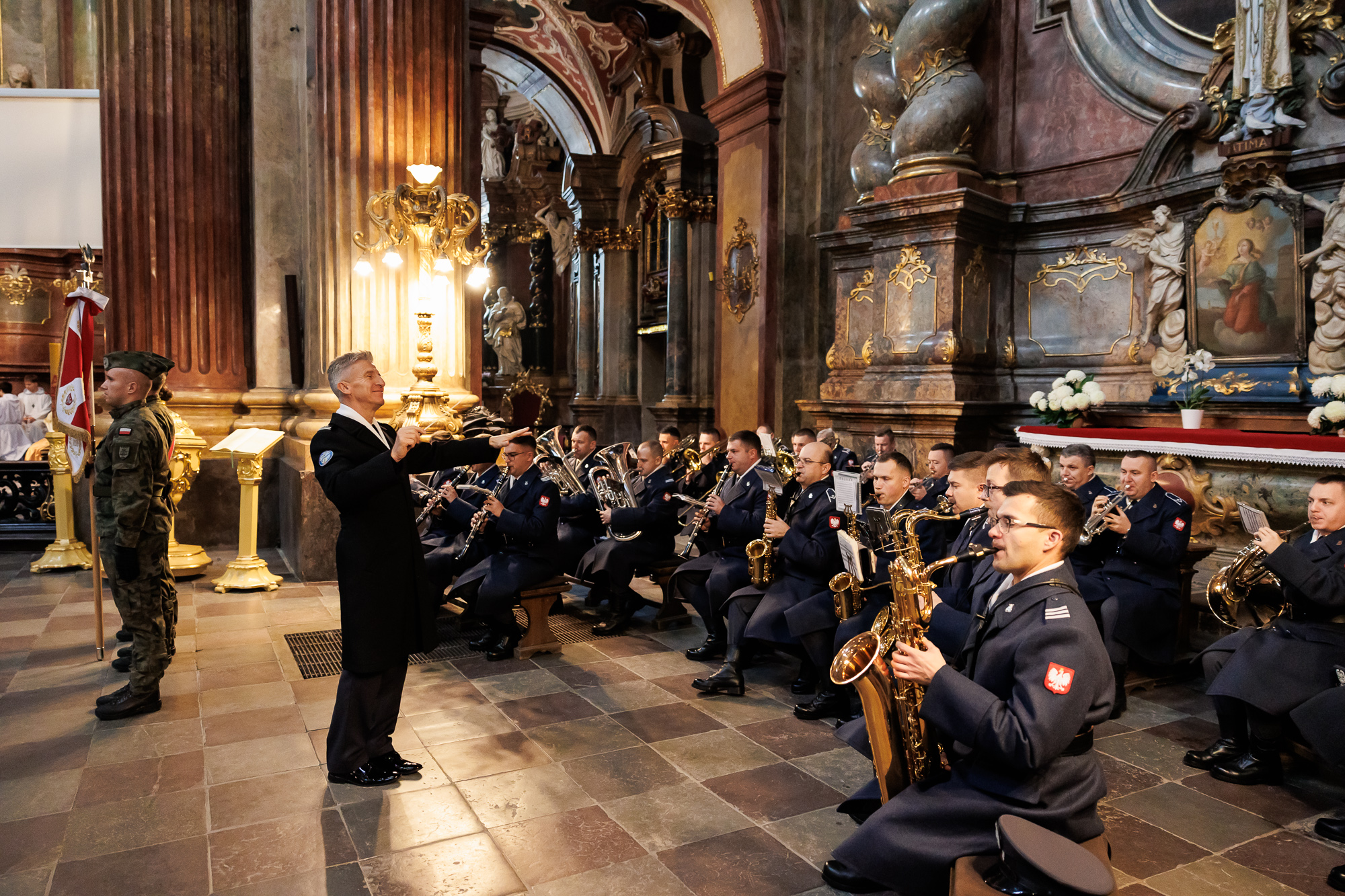 Rząd muzyków w wojskowych mundurach siedzi z instrumentami w kościele, a przed nimi stoi dyrygent.