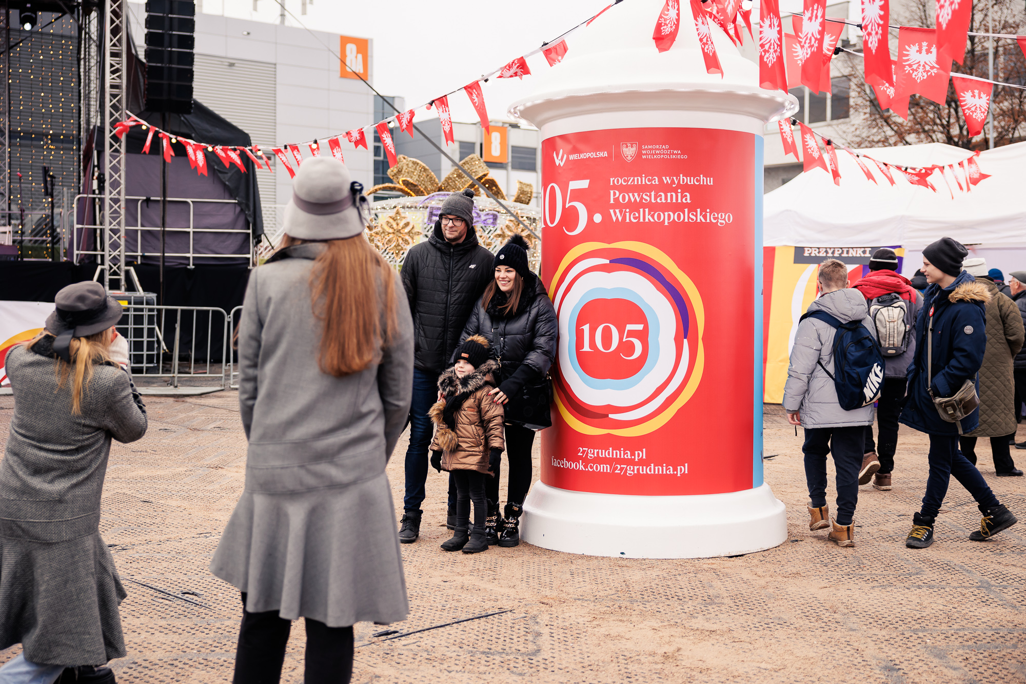 Słup reklamowy z plakatem reklamującym 105. rocznicę wybuchu Powstania Wielkopolskiego, przy którym stoi mężczyzna z kobietą i małym dzieckiem, przygląda się temu dziewczyna ubrana w ubiór damski z czasów powstania, stojąca tyłem od patrzącego.