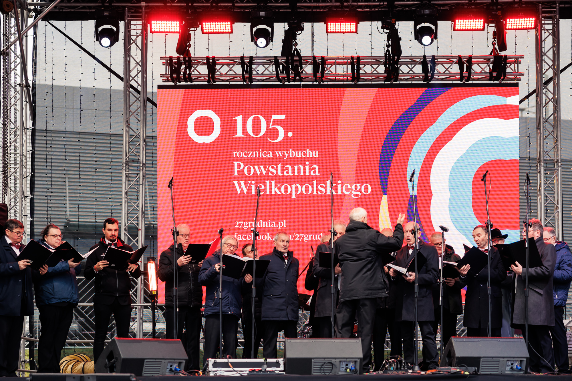 Grupa osób zebranych na scenie koncertowej, śpiewających wspólnie z trzymanych nut.