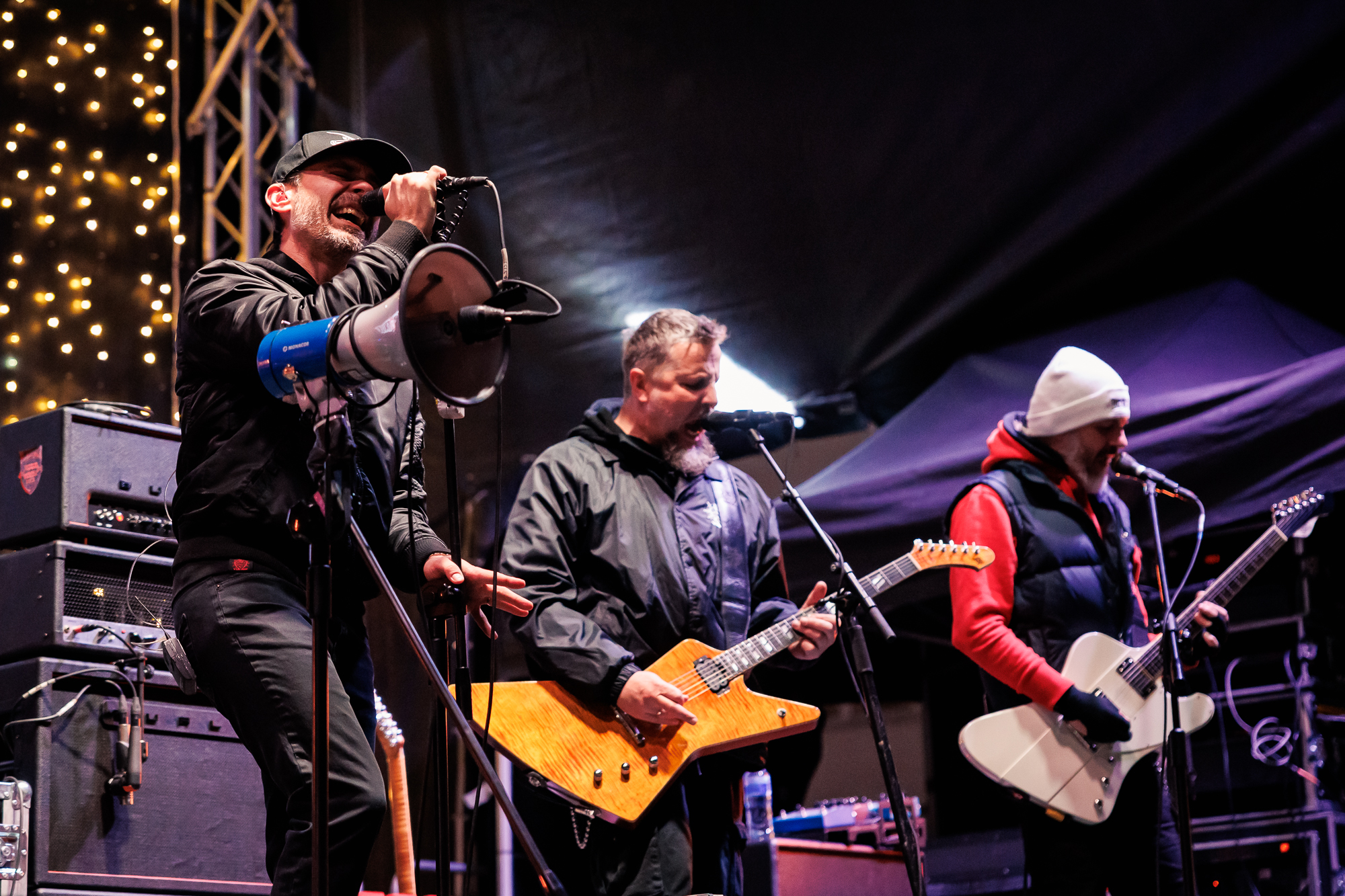 Gitarzyści na scenie, obok mężczyzna śpiewający do mikrofonu.