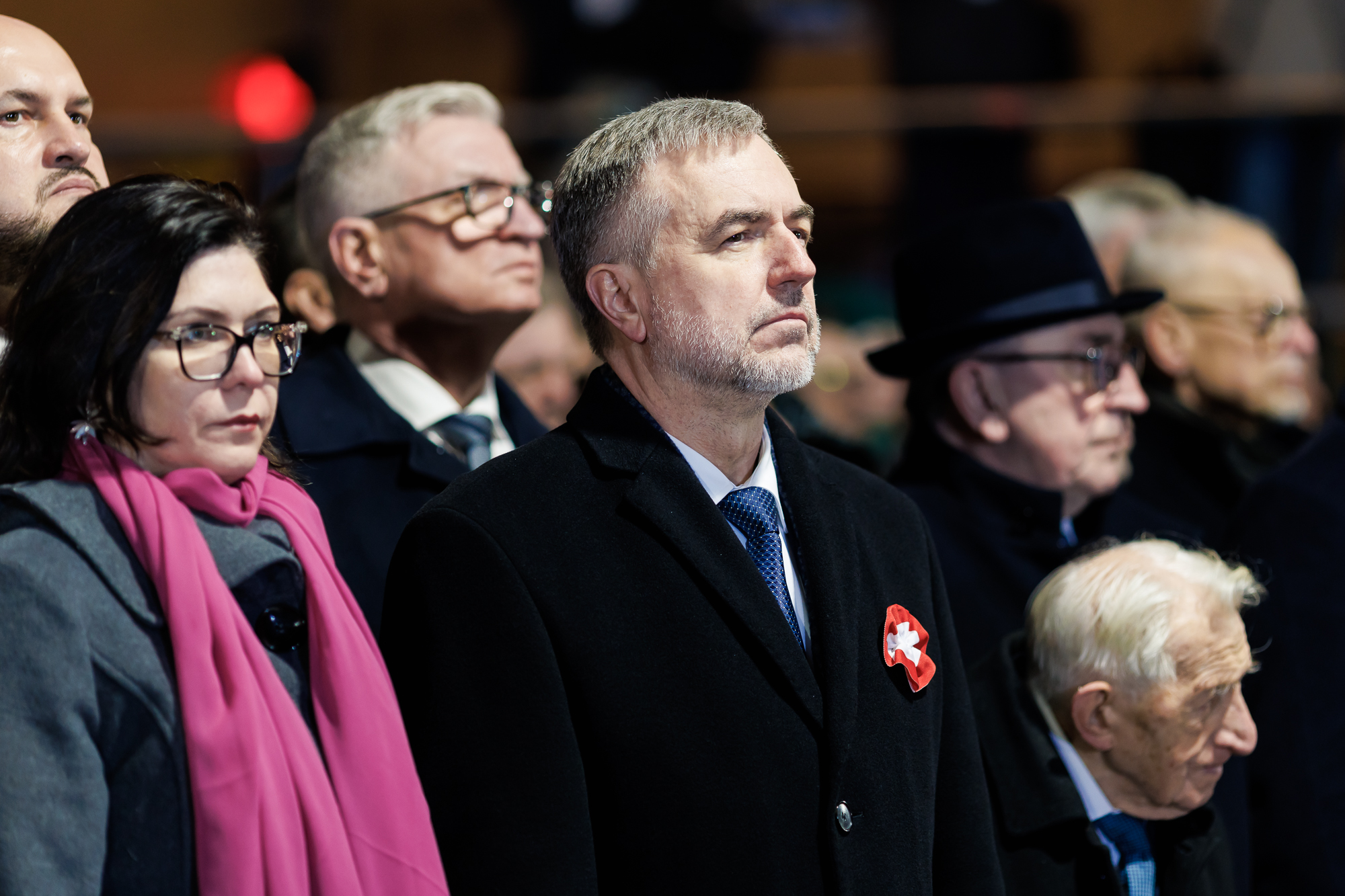 Mężczyzna w płaszczu z rozetką powstańczą w klapie spogląda przed siebie. Za nim widać głowę drugiego mężczyzny w okularach i płaszczu.