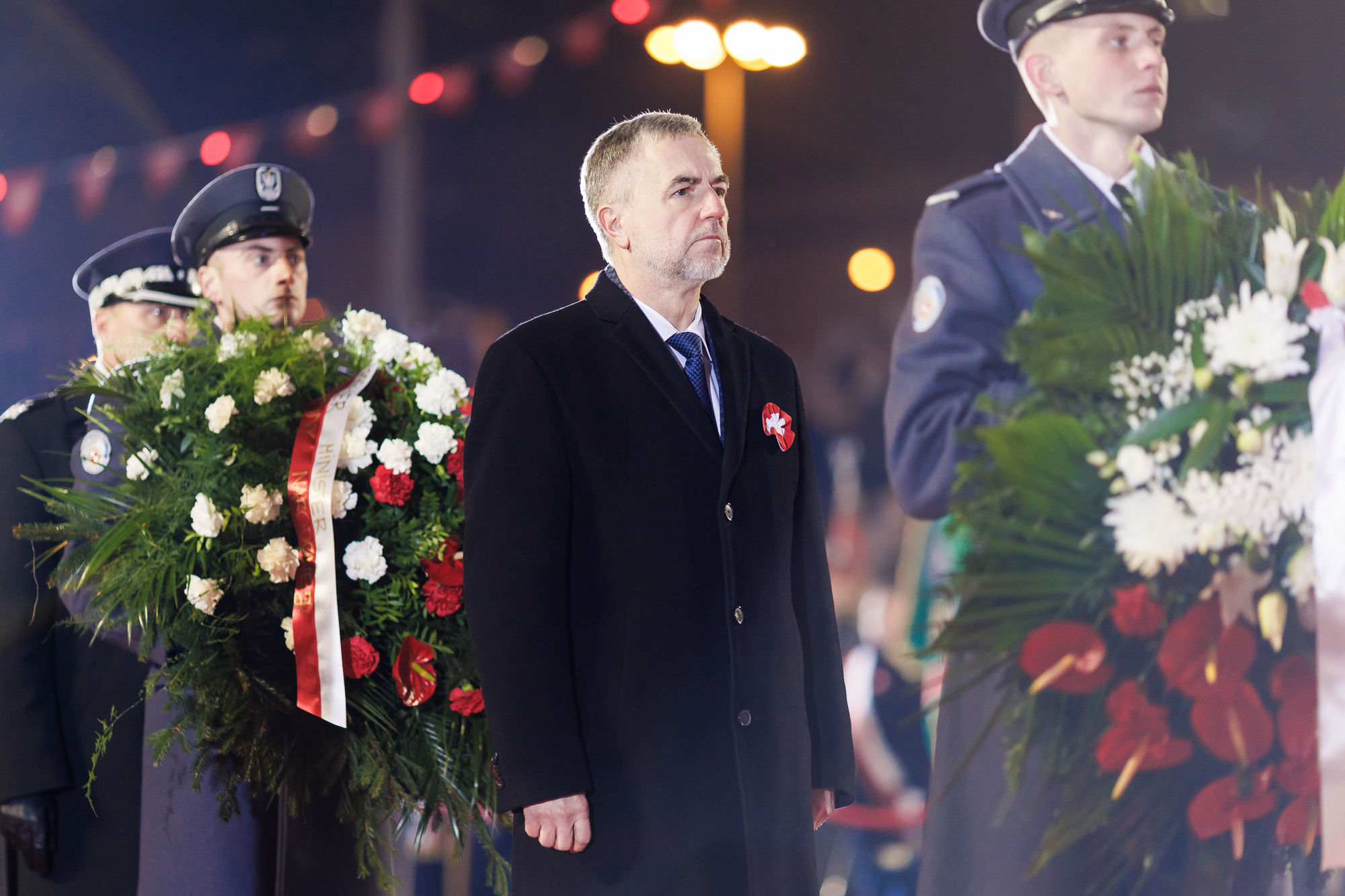 Mężczyzna w czarnym płaszczu z rozetą powstańczą stoi za wojskowym w granatowym płaszczu i wiązanką w rękach, za mężczyzną w ciemnym płaszczu stoi kolejny wojskowy w błękitnym płaszczu z wiązanką w rękach.