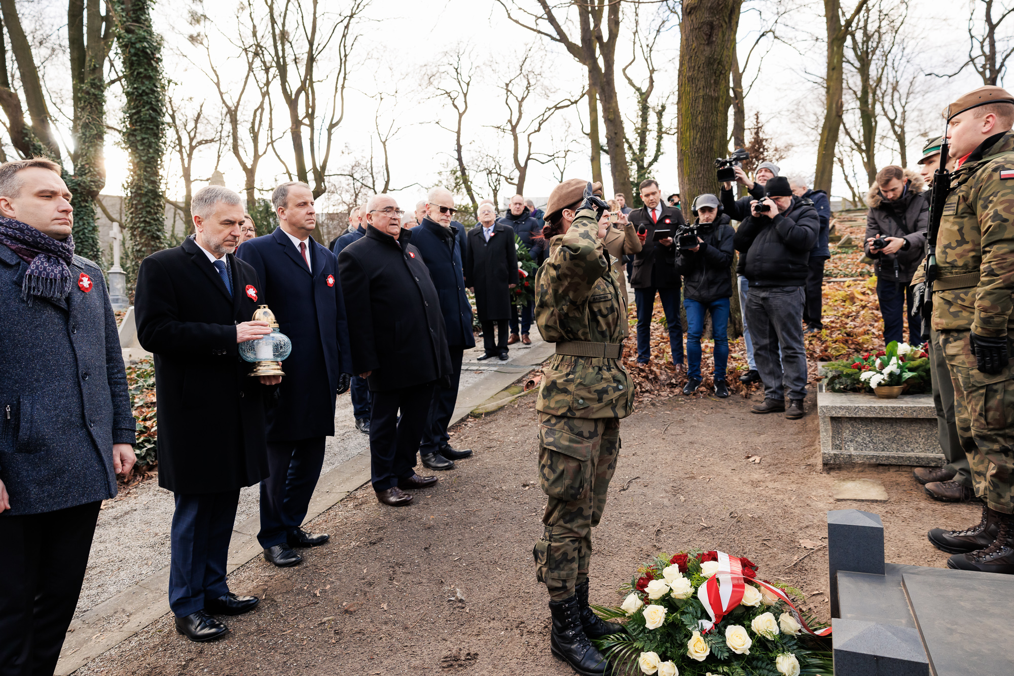 Żołnierka salutująca, przy jej nogach wiązanka. Za jej plecami rząd mężczyzn, jeden z nich trzyma zapalony znicz.