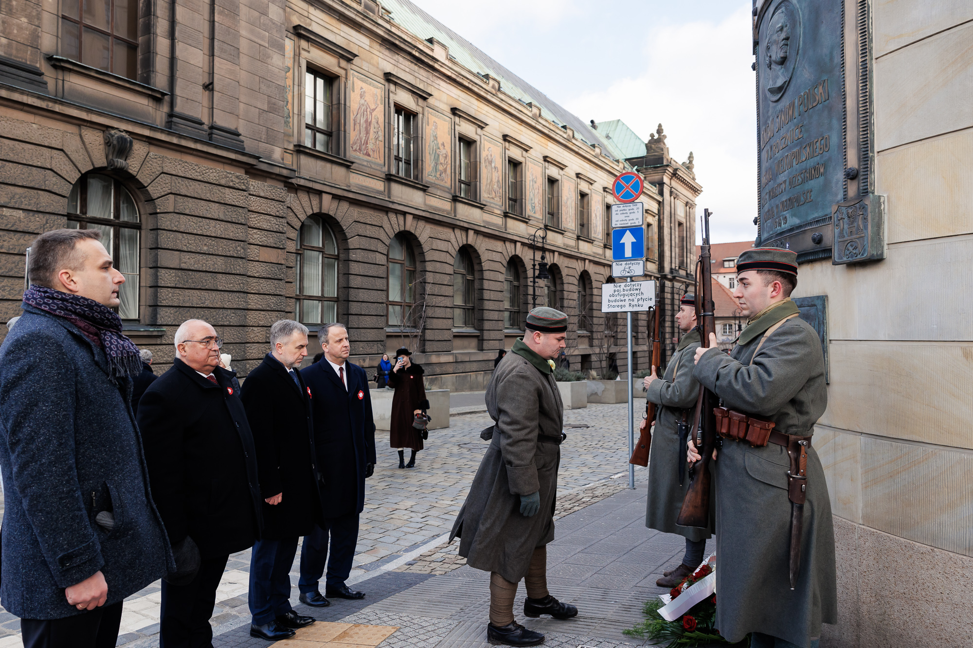 Osoba w mundurze powstańczym składa kwiaty przy tablicy zwieszonej na narożniku budynku, a za jego plecami stoją mężczyźni.. W tle brukowana ulica i budynek.