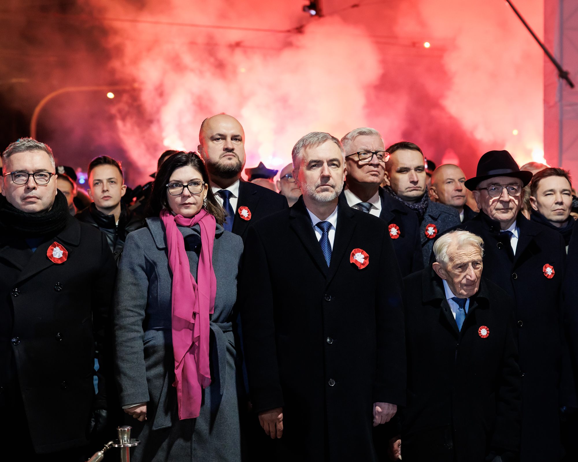 Tłum ludzi stojącyna wprost do patrzącego, w tle czerwone światła i dymy z rac.