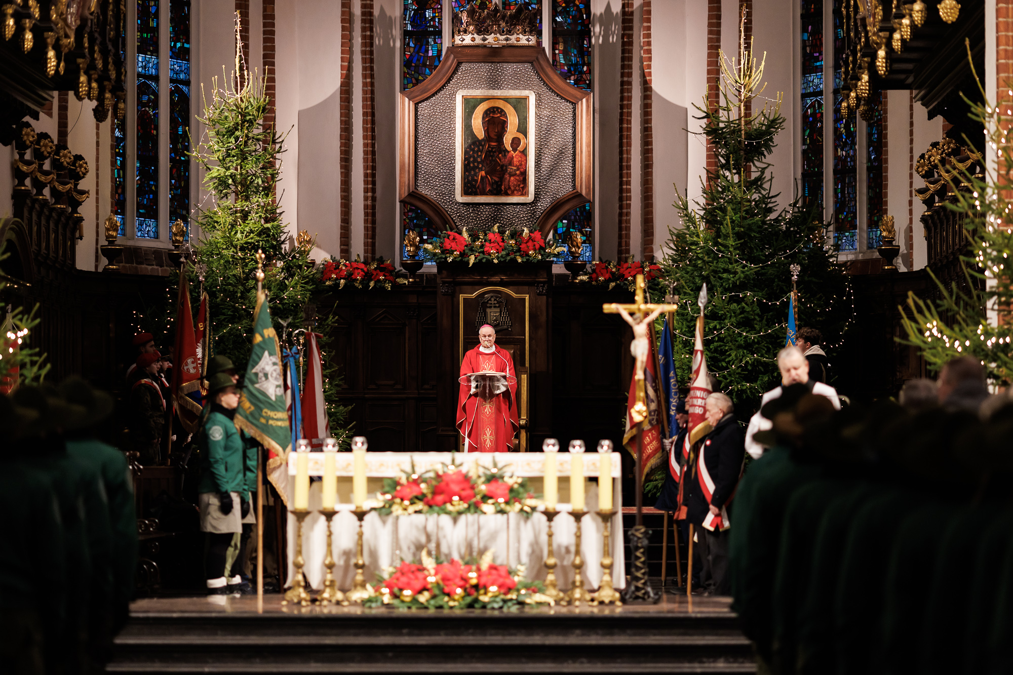 Widok na ołtarz w kościele, w tle obraz Matki Boskiej Częstochowskiej. Przy ołtarzu w czerwonym ornacie stoi ksiądz.