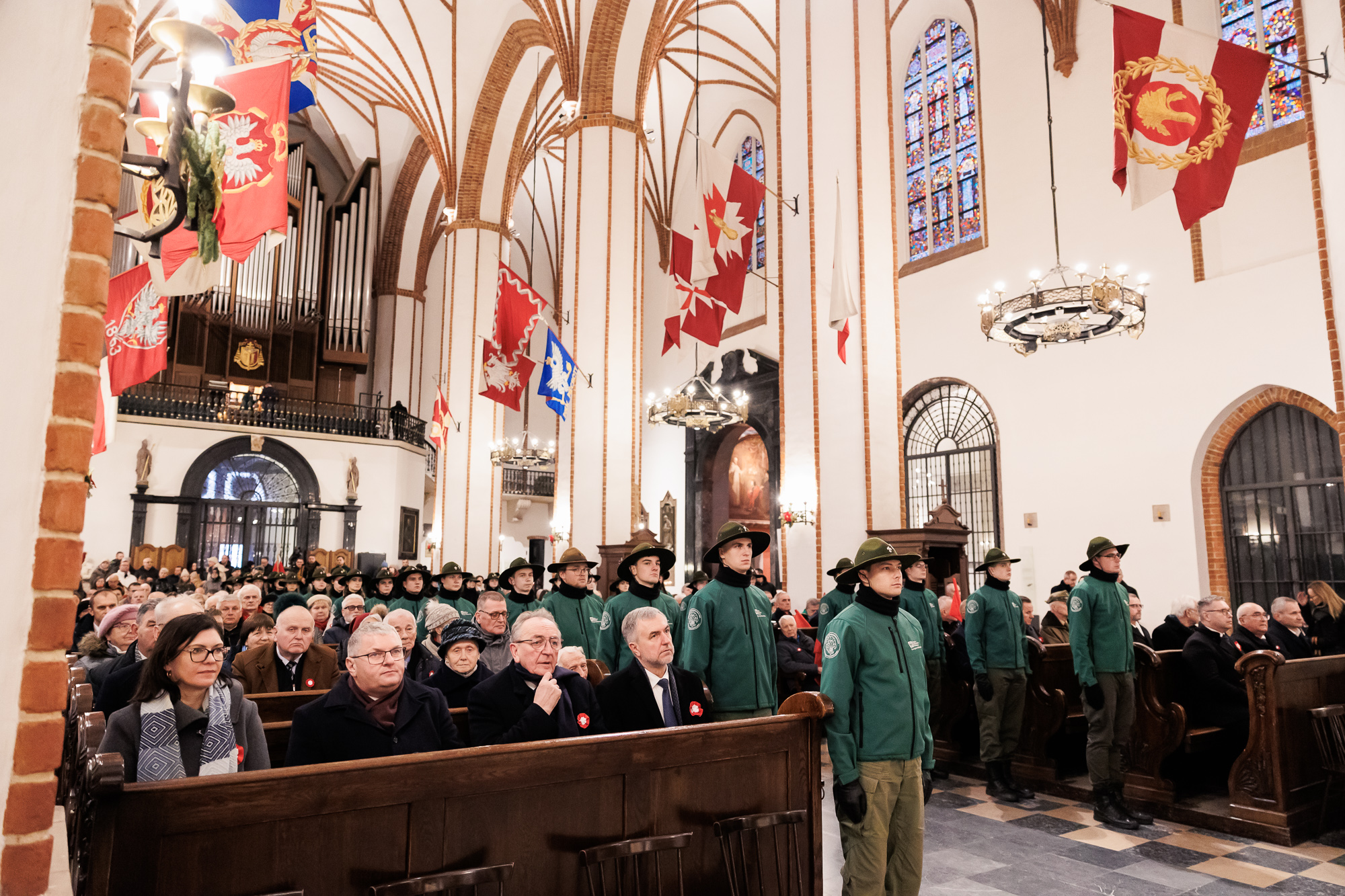 Widok na wnętrze kościoła. Na pierwszym planie siedzą trzej mężczyźni w ciemnych płaszczach. Obok każdego rzędu łąwek stoi harcerze w zielonym mundurze i szerokim kapeluszu.