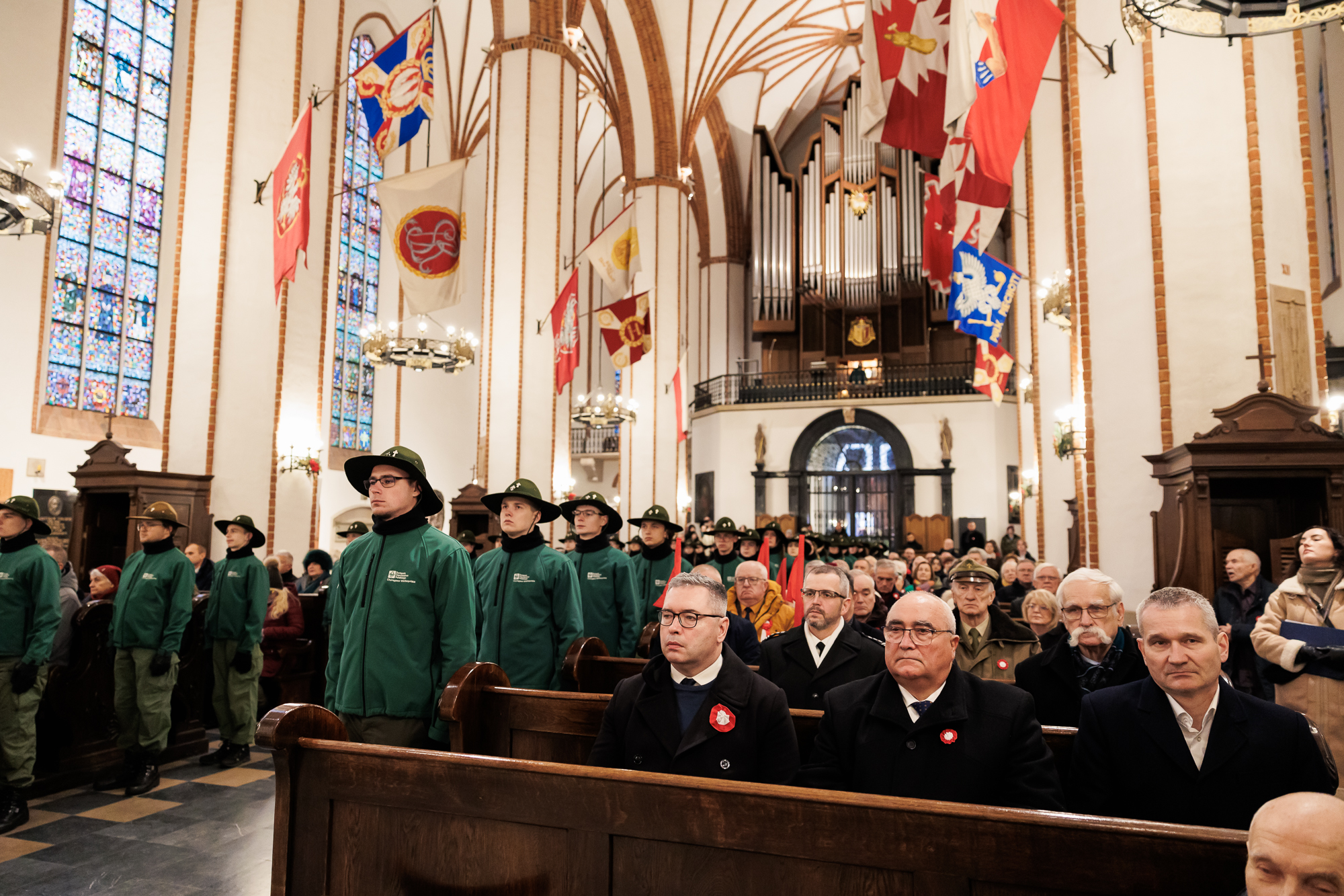 Widok na wnętrze kościoła. Na pierwszym planie siedzą dwaj mężczyźni w ciemnych płaszczach. Obok każdego rzędu łąwek stoi harcerze w zielonym mundurze i szerokim kapeluszu.