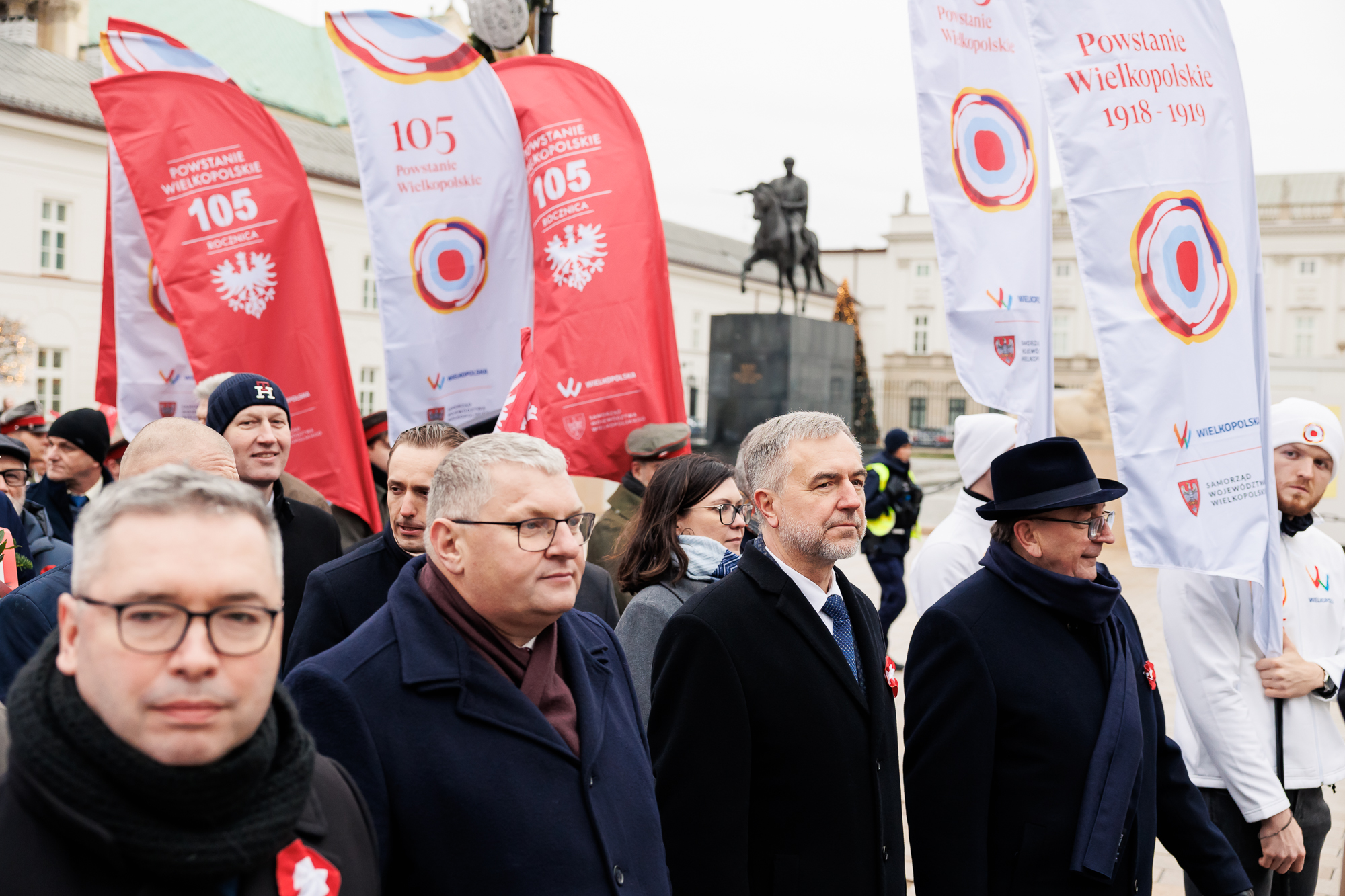 Trzej mężczyźni w ciemnych płaszczach idą na czele pochodu. Za ich plecami widać tłum niosący banery pionowe z logotypem rozety powstańczej i orła.