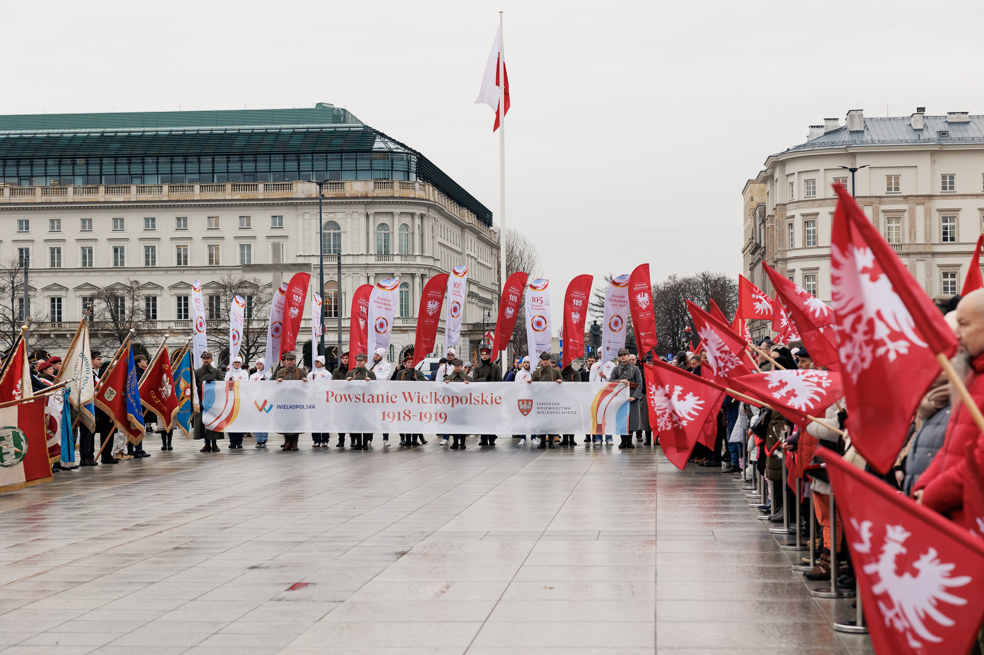 Czoło pochodu wchodzi na plac. Na czele widoczny poziomy baner z napsiem 105. rocznica wybuchu Powstania Wielkopolskiego. W tle po lewej stronie widać duży budynek dwupiętrowy zielonym dachem.