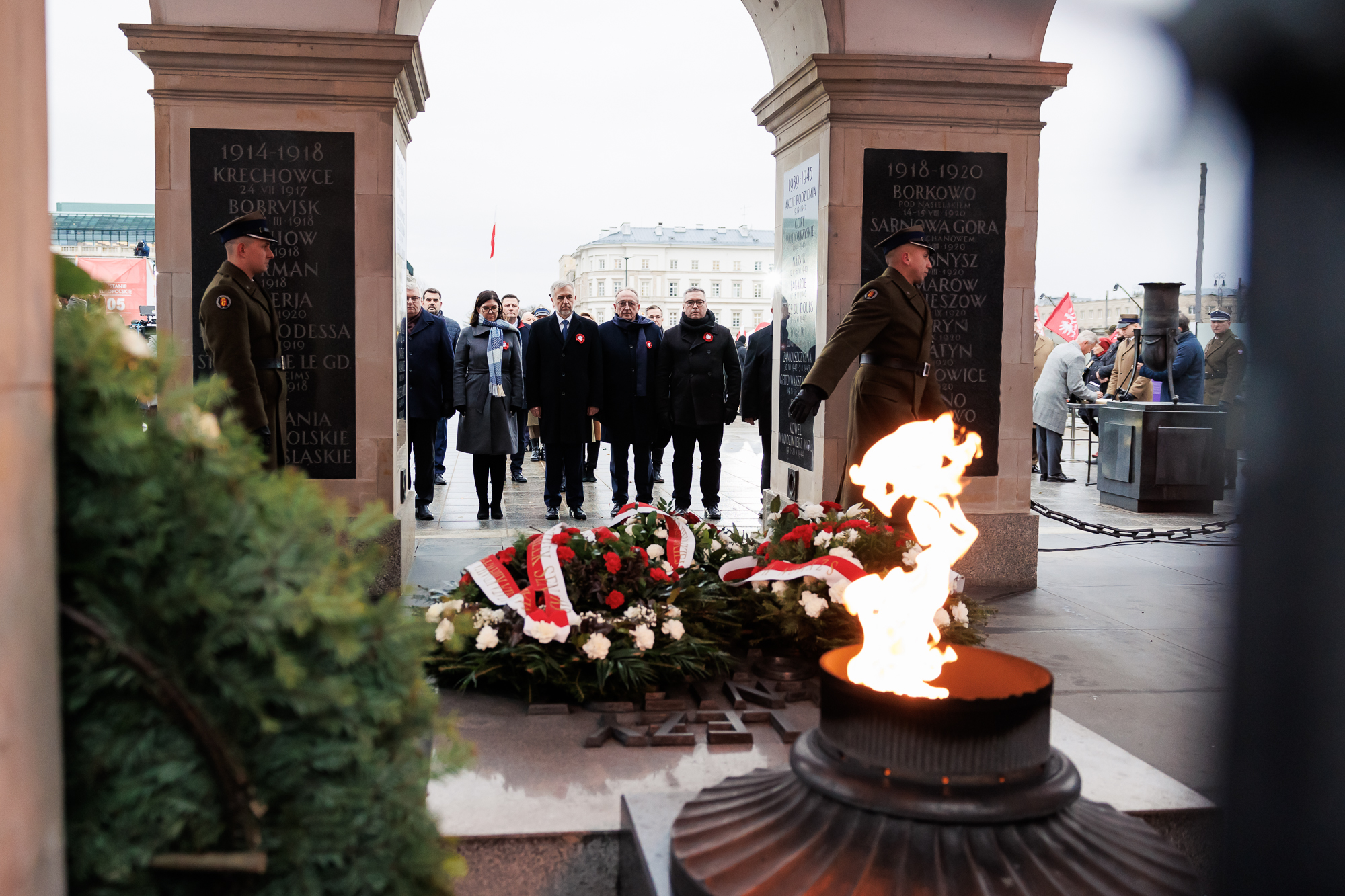 Na pierwszym planie widoczny płonący ozdobny znicz i płyta nagrobna z leżącą na niej wiązanką. W tle stojacy pod filarami Grobu Nieznanego Żołnierza ludzie w rzędzie z kwiatami.