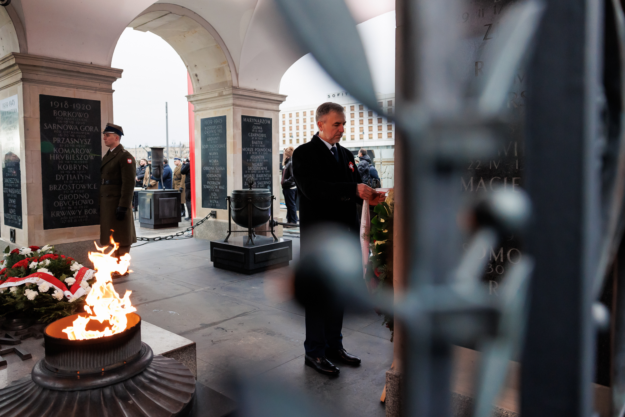 Mężczyzna stoi sklepieniem z filarami Grobu Nieznanego Żołnierza. W tle widoczny filar z tablicami oraz stojący żołnierz