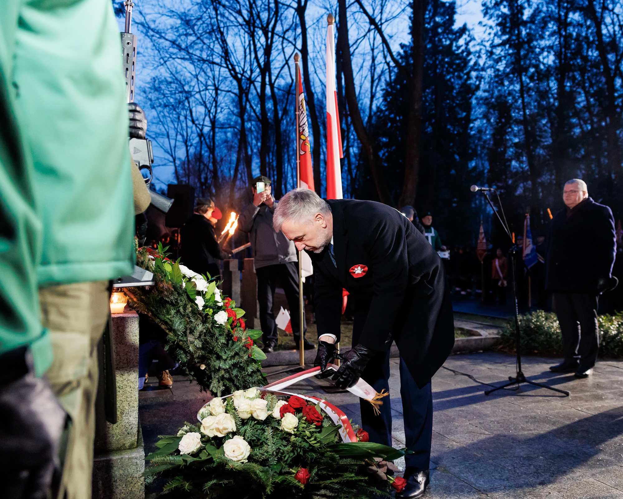 Mężczyzna skłąda wieneic przy pomniku. W tle drzewa i zapadający wieczór.