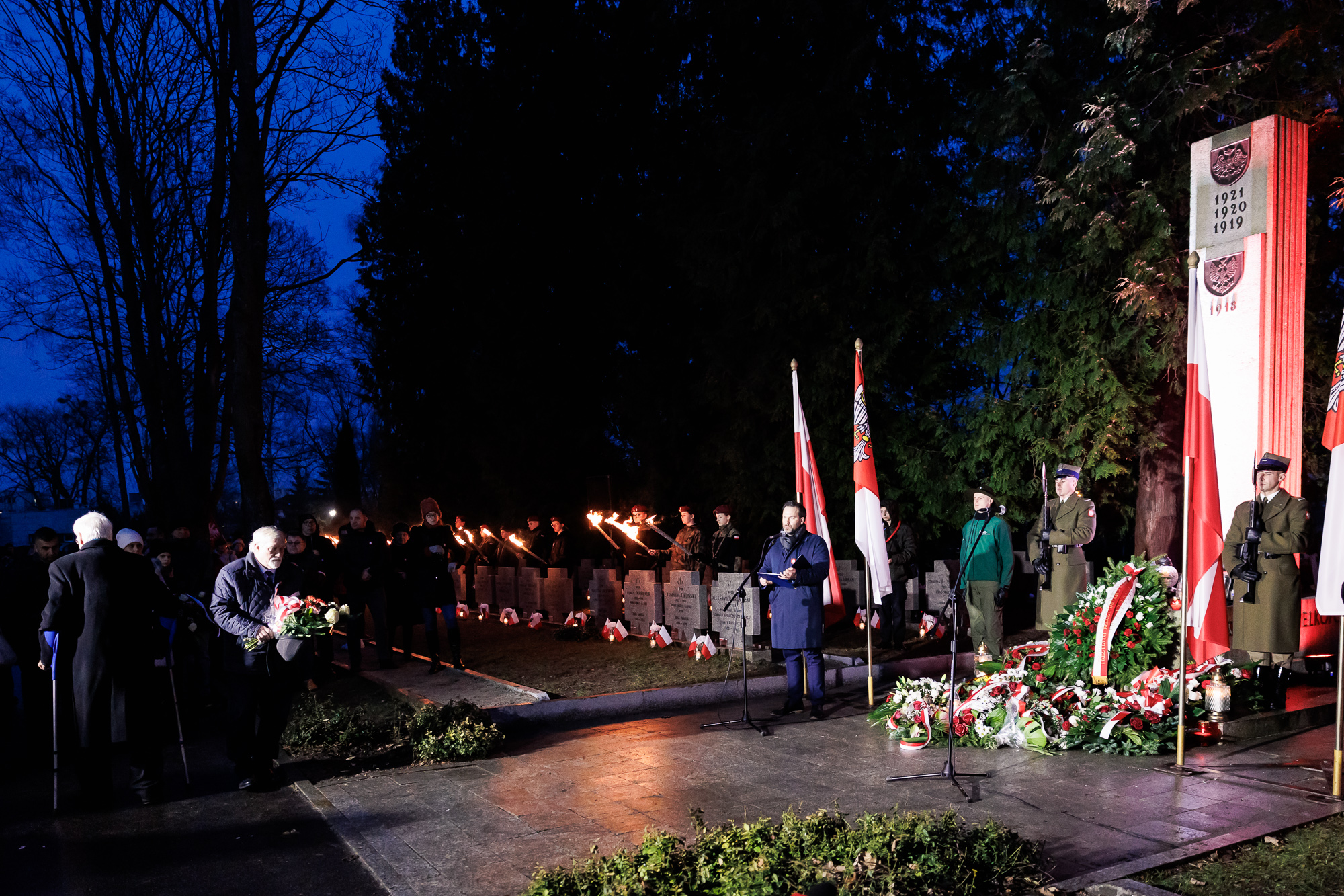 Widok na fragment kwaitów i wieńcy przed pomnikiem. W tle przy mikrofonie stojący mężczyzna. Po lewej stronie mężczyzna idący złożyć kwiaty.