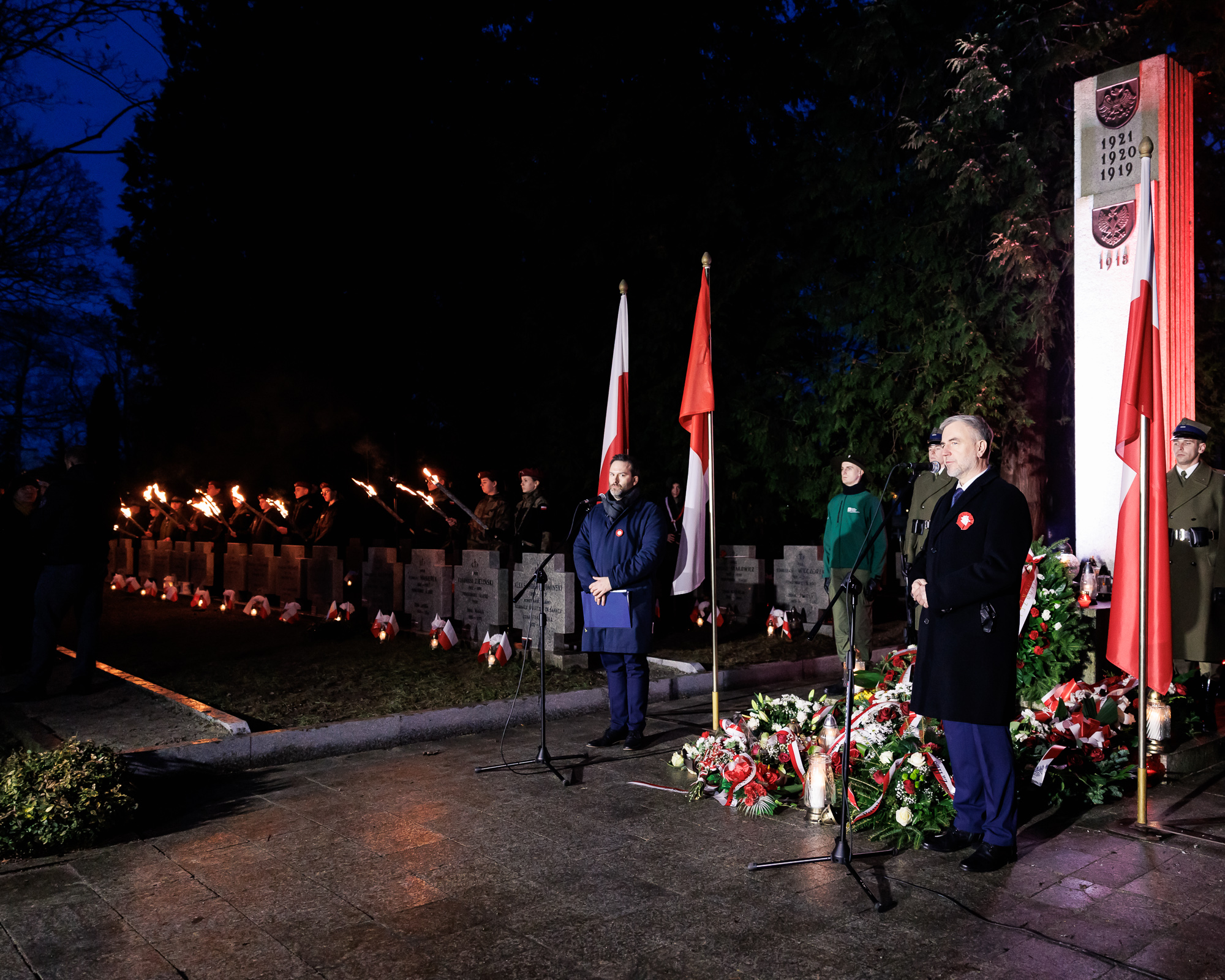 Mężczyzna przemawia do mikrofonu na tle pomnika i kwiatów. W tle stojący z płonącymi pochodniami harcerze.