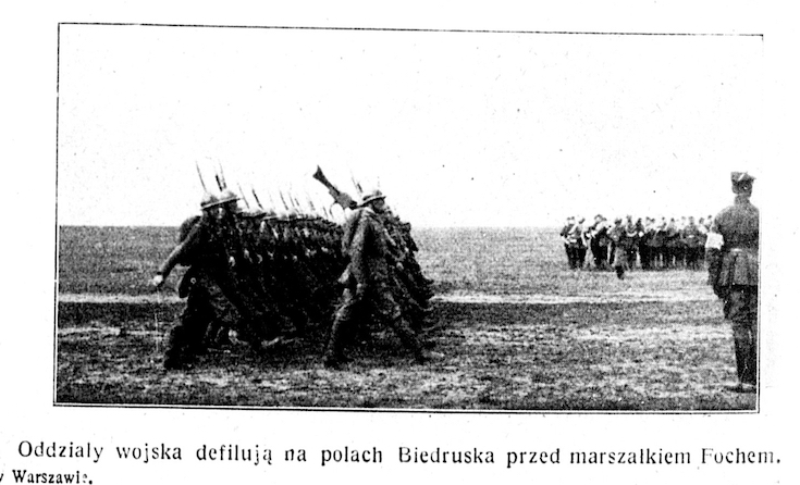Wielu żołnierzy w mundurach, część z nich stoi, a pozostali maszerują z bronią na ramieniu.