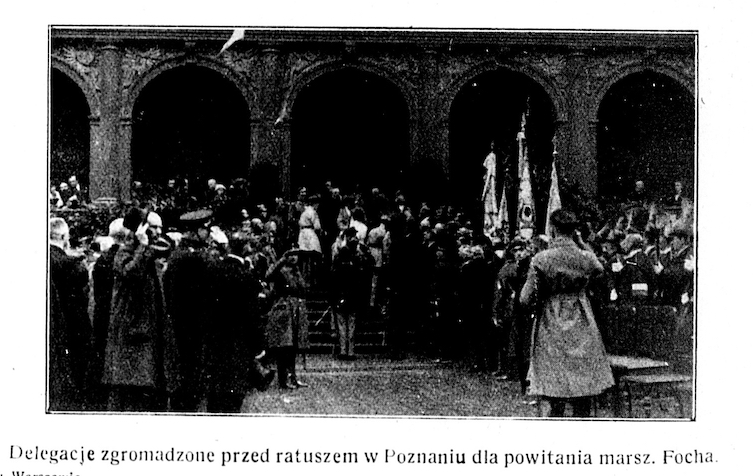 Tłum ludzi stojących przed budynkiem, widać też kilka sztandarów.