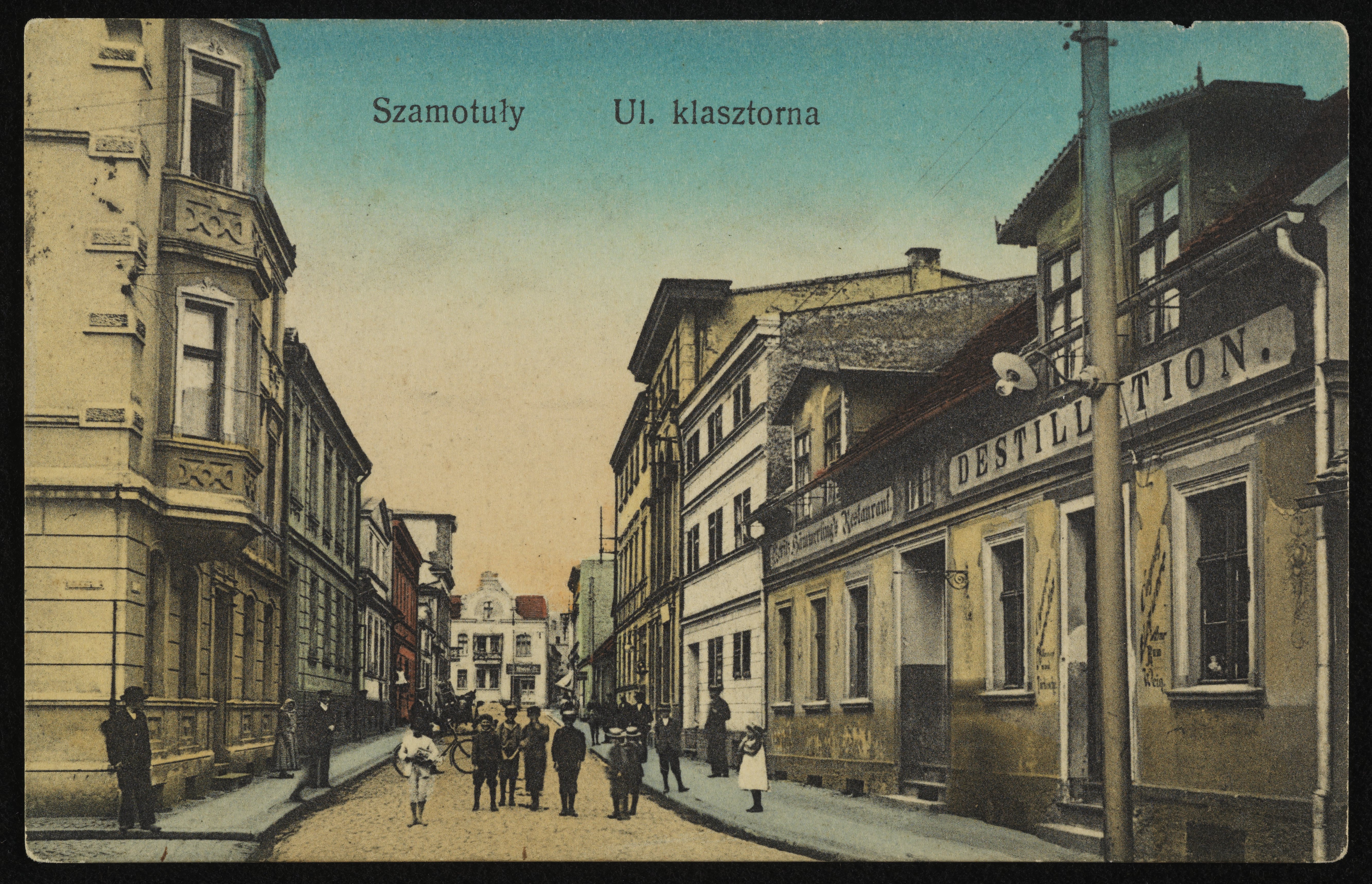 Dzieci stojące na ulicy. Po obu jej stronach stoją domy i kamienice.