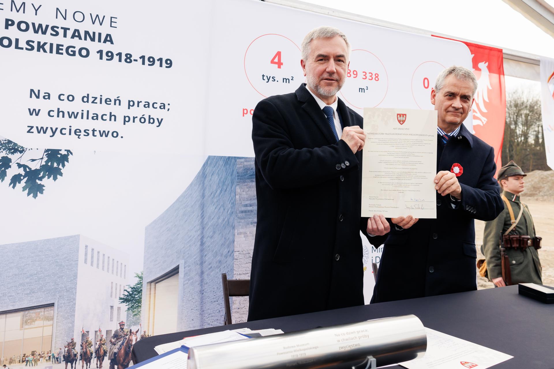 Dwóch mężczyzn w czarnych płaszczach trzyma razem kartkę papieru. W tle tablica informacyjna i mężczyzna w mundurze.