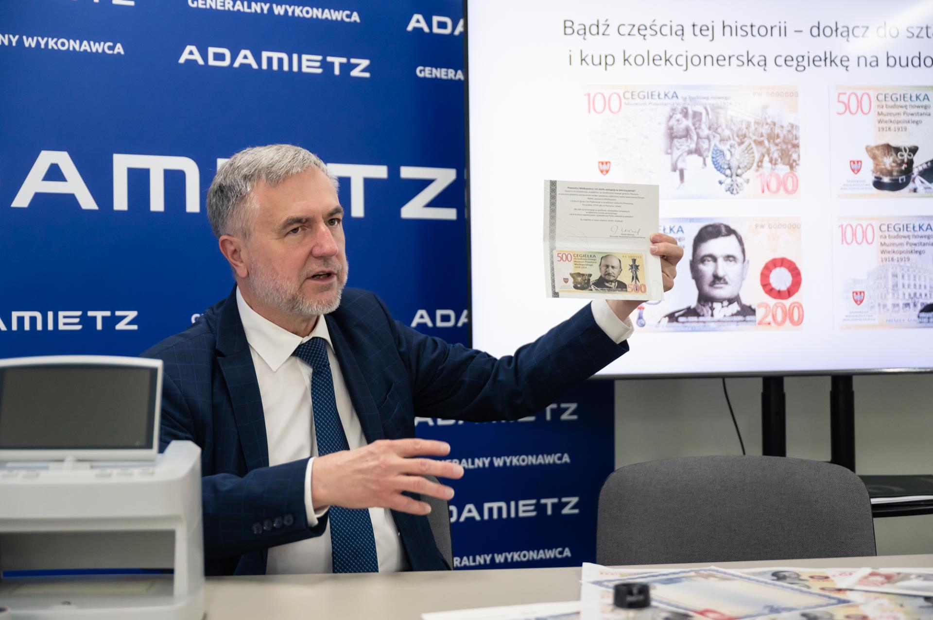 Mężczyzna siedzi za stołem, pokazuje papierową zadrukowaną kartkę – okolicznościową cegiełkę, w tle tablice informacyjne.