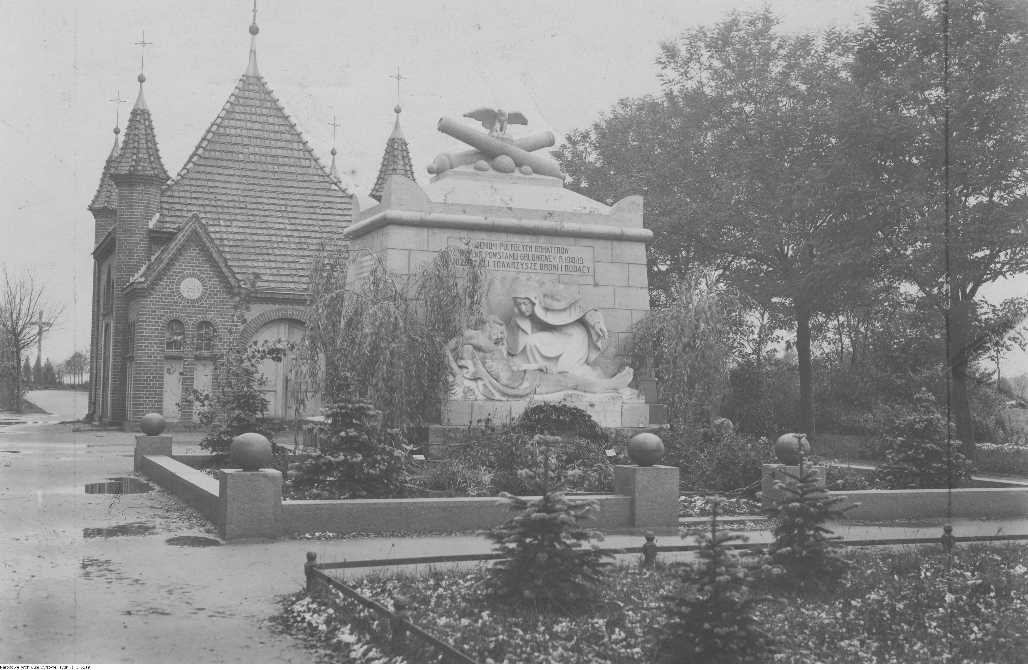 Pomnik na cmentarzu, w jego centrum postać klęczącej kobiety, w tle budynek kaplicy