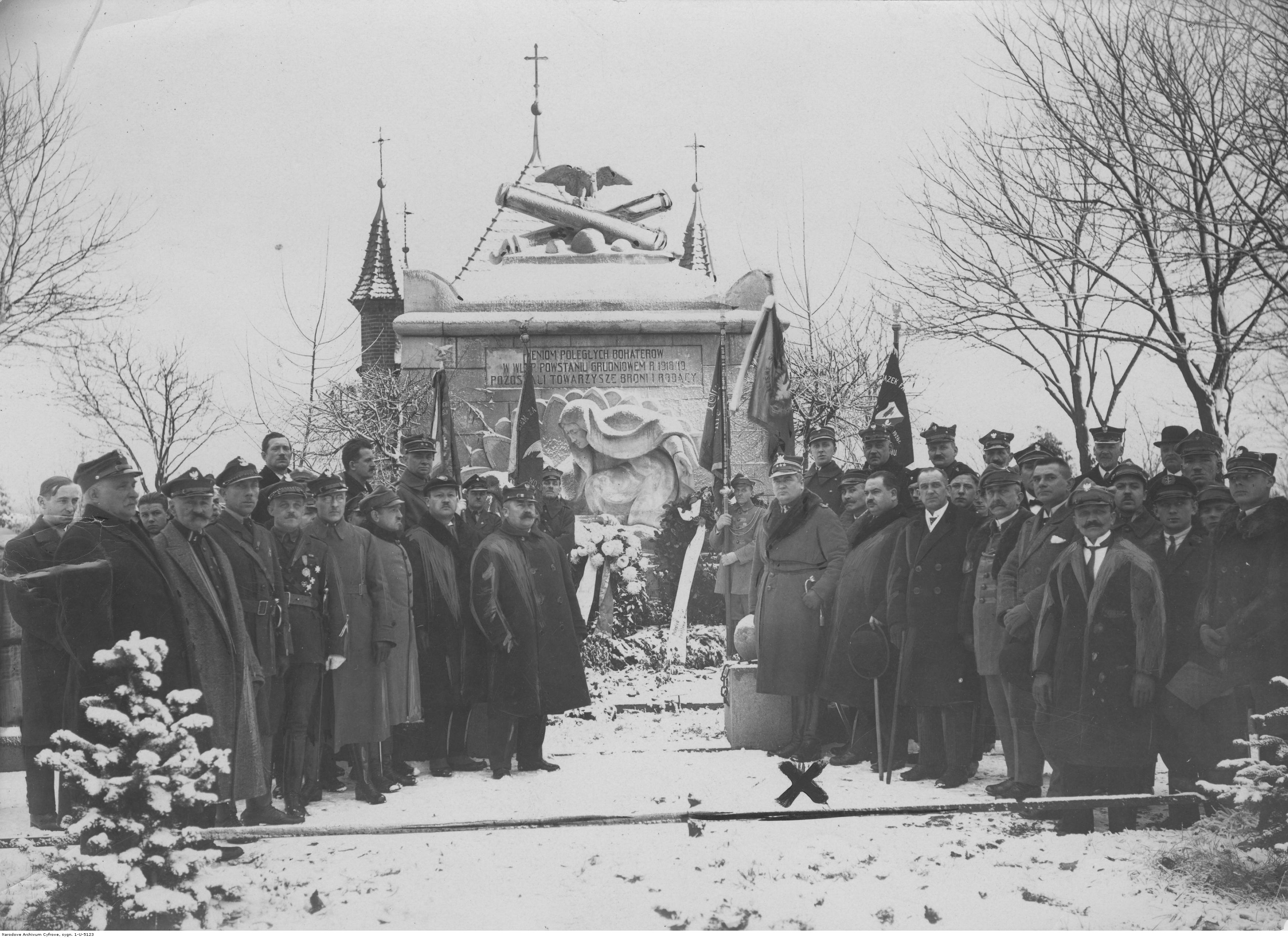Grupa mężczyzn przed pomnikiem, część z nich w mundurach, pomnik pokryty śniegiem