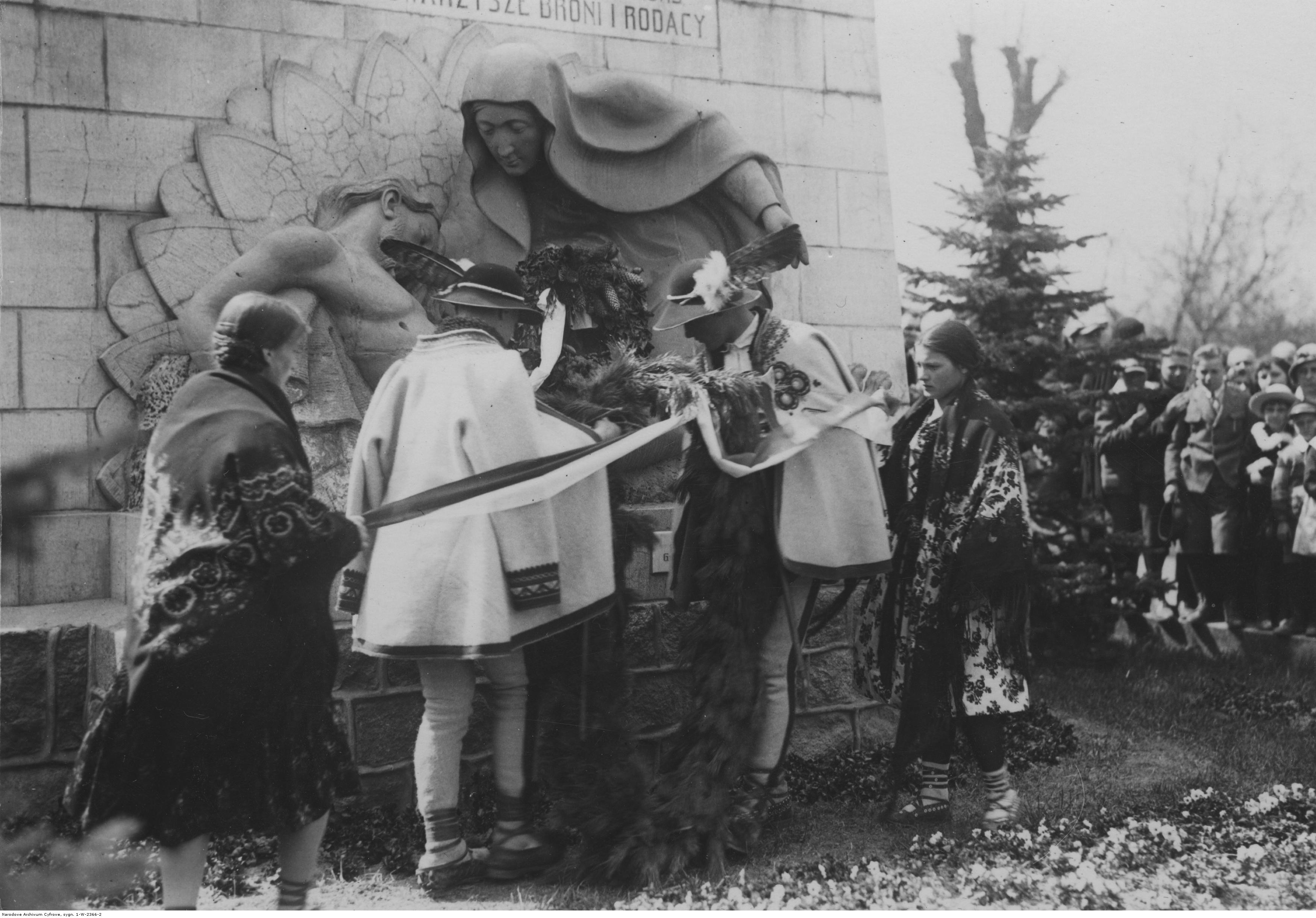 Kobiet i mężczyźni w strojach ludowych, składają kwiaty pod pomnikiem