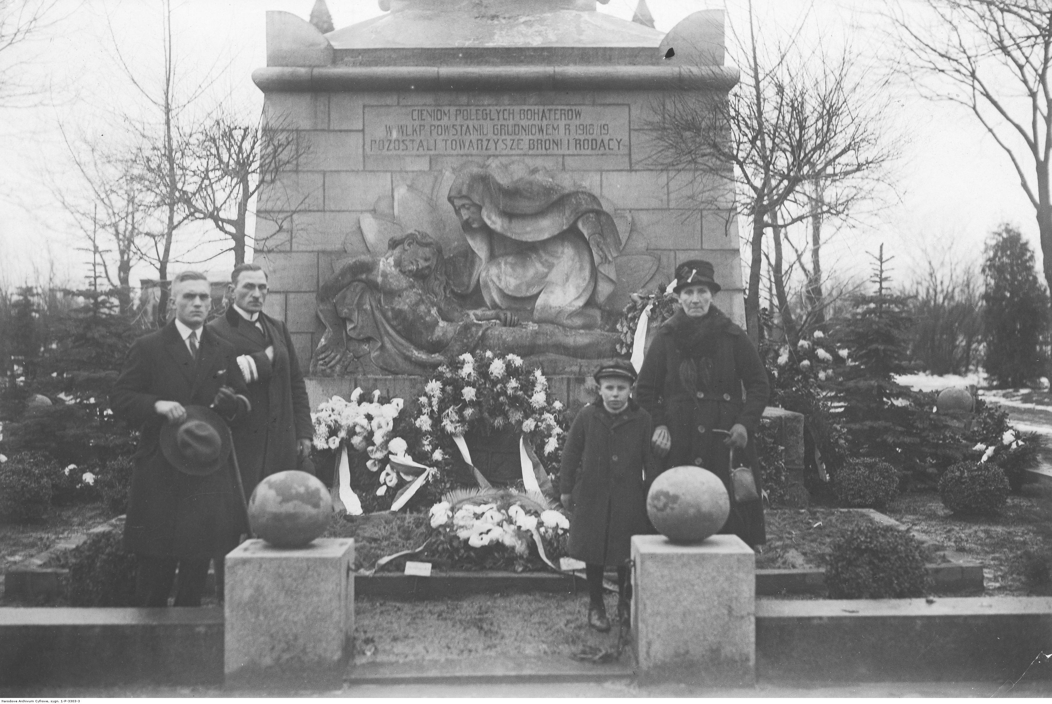 Grupa ludzi w różnym wieku przed pomnikiem, w tle wiązanki kwiatów