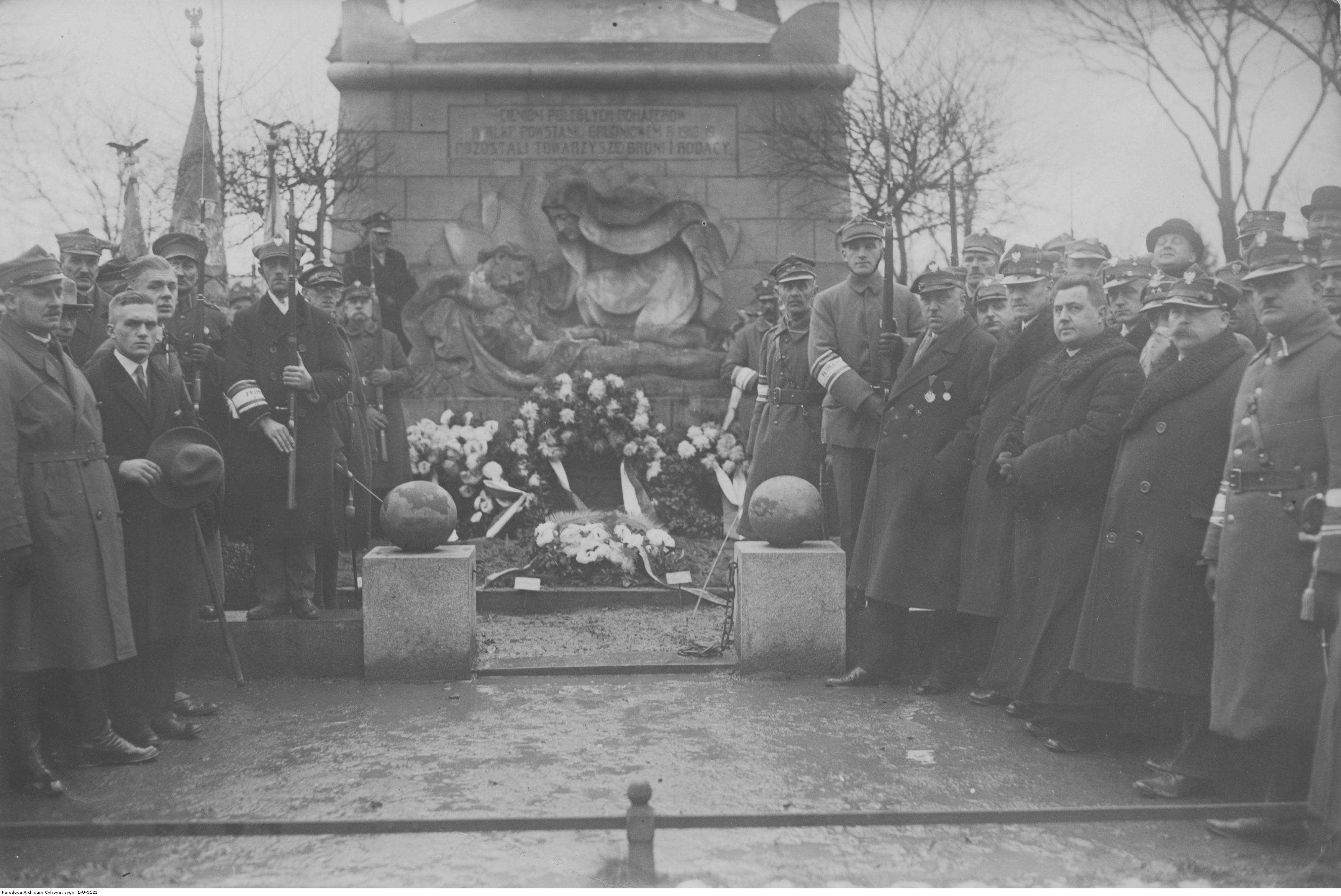 Grupa mężczyzn przed pomnikiem, wielu z nich w mundurach wojskowych, część prezentuje broń