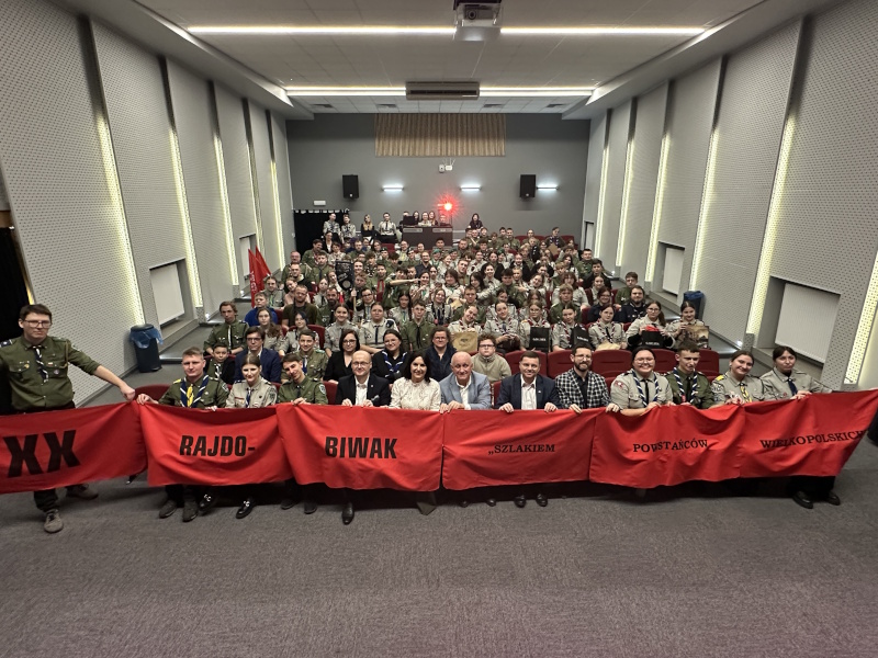 Na zdjęciu duża grupa ludzi, uczestników spotkania. Osoby w pierwszym rzędzie trzymają czerwony baner z nazwą wydarzenia