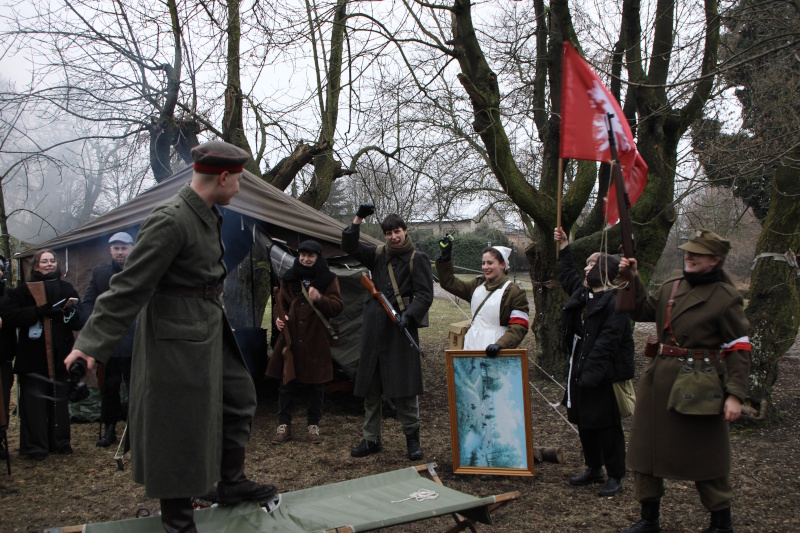 Uczestnicy zadania terenowego stoją ubrani w stroje z epoki. W centralnej części dziewczyna w stroju sanitariuszki trzyma obraz. Jeden z uczestników gry terenowej trzyma powstańczą flagę. Pozostali uczestnicy są ubrani w stroje z epoki.