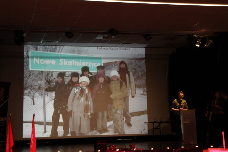 Na scenie, na ekranie zdjęcie z wydarzenia, uczestnicy stoją na tle tablicy z nazwą Nowe Skalmierzyce