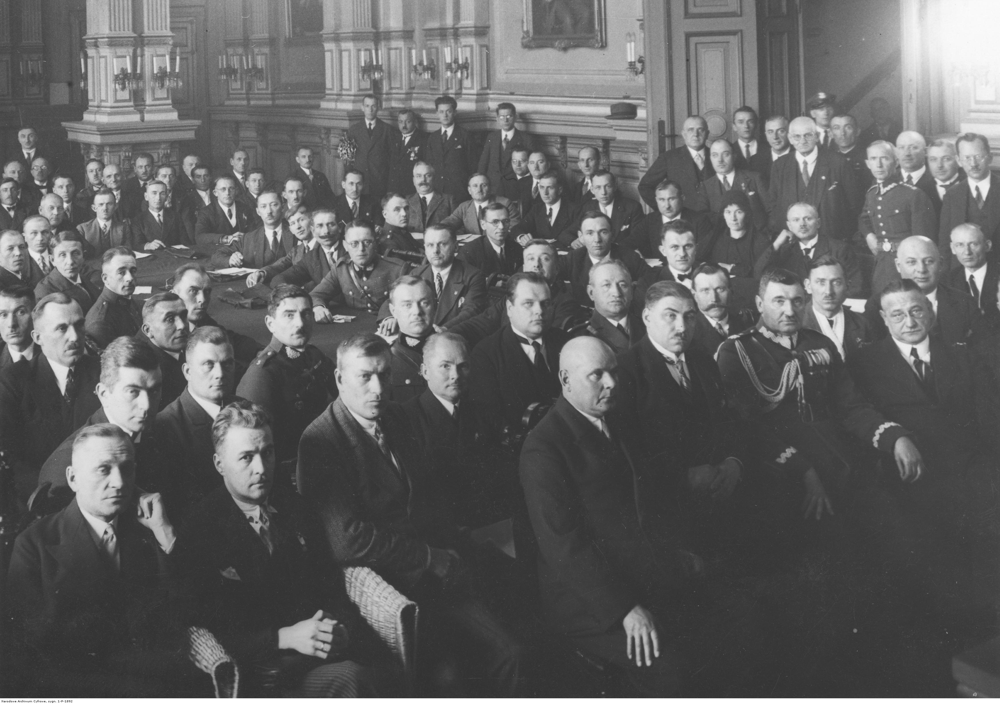 Duża grupa mężczyzn w odświętnych strojach, siedzą w dużej sali, część z nich  w mundurach.