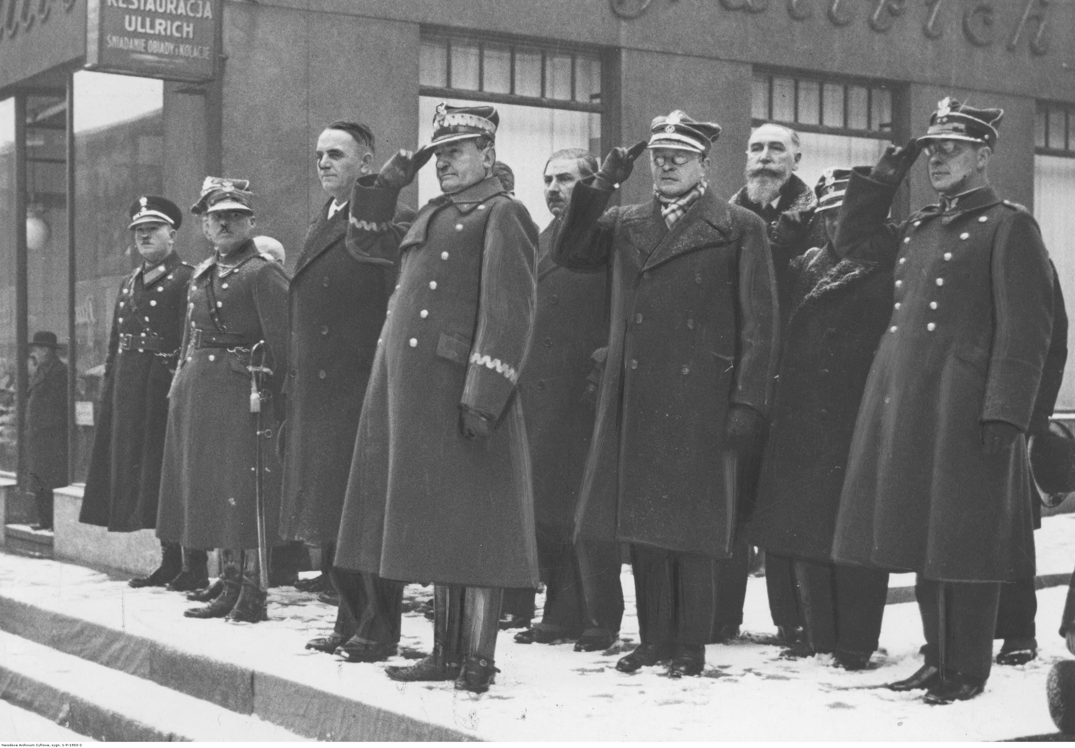 Grupa mężczyzn przed budynkiem, część z nich salutuje.
