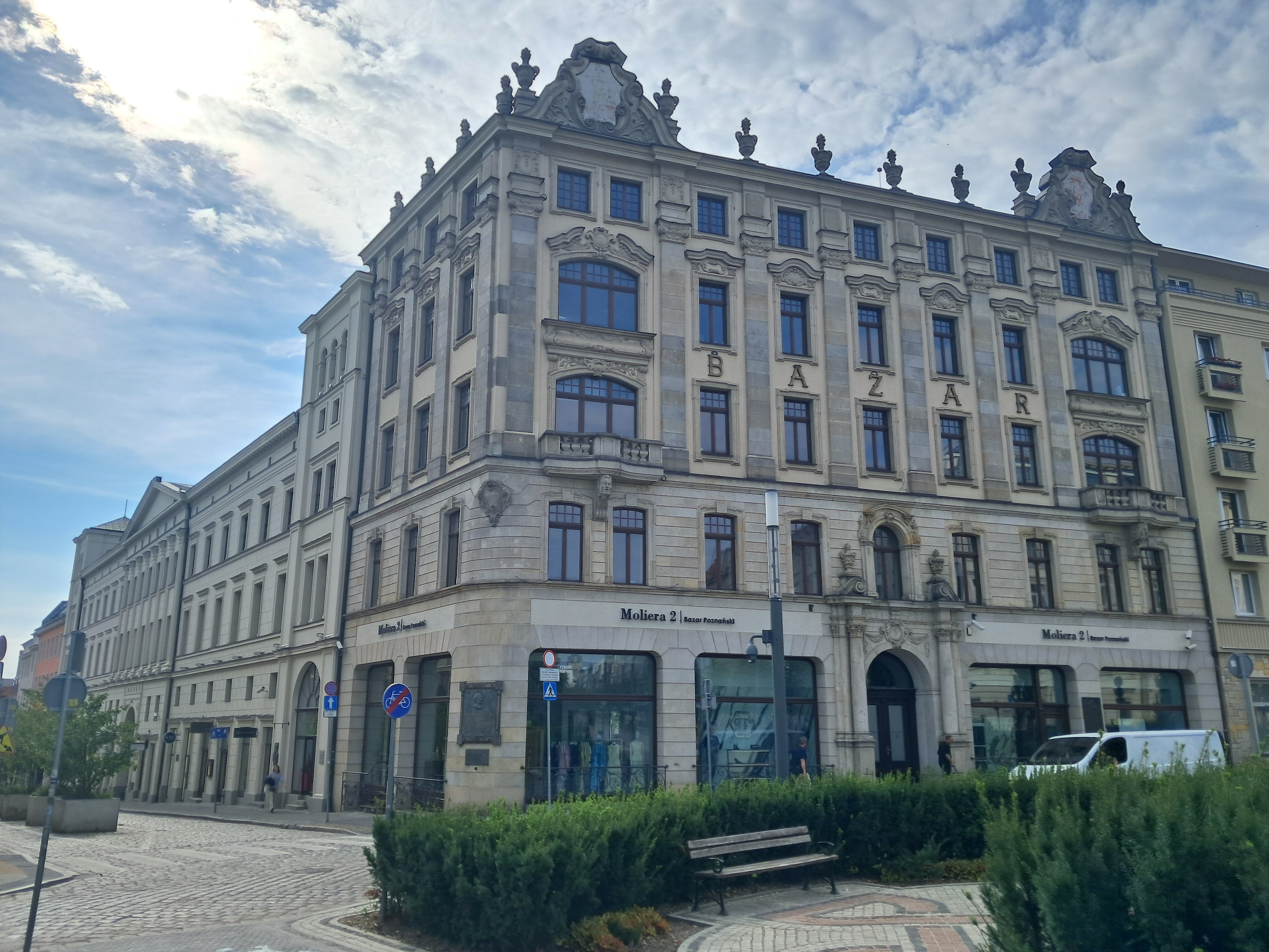 Współczesny widok dużego, zabytkowego budynku z napisem „BAZAR”; na parterze sklepy, przed nimi przechodnie i biały samochód.